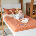 Smiling young boy drawing on a notebook in a cozy modern kids bedroom, lying on an orange quilted bed with a wooden bunk-frame, markers scattered and a dinosaur plush toy nearby