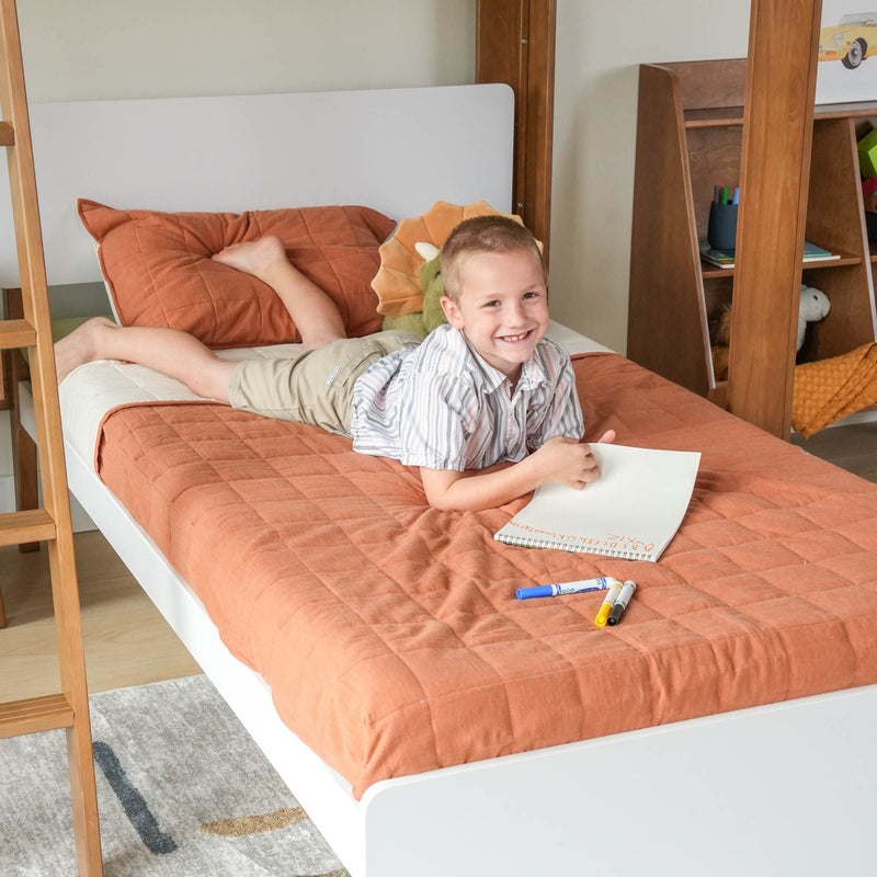 Smiling young boy drawing on a notebook in a cozy modern kids bedroom, lying on an orange quilted bed with a wooden bunk-frame, markers scattered and a dinosaur plush toy nearby