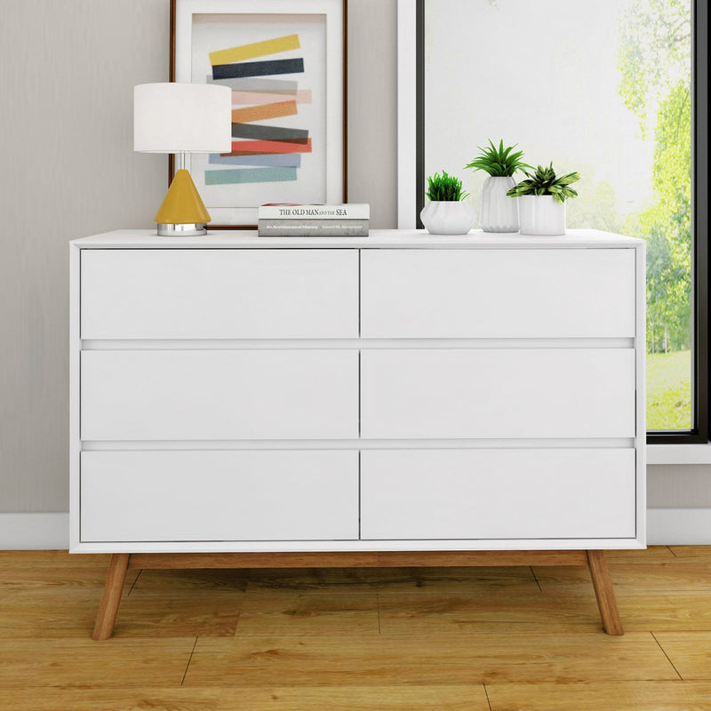 Modern white dresser with six drawers, wooden legs, and decorative items including a lamp, books, and small plants, set against a neutral wall background.