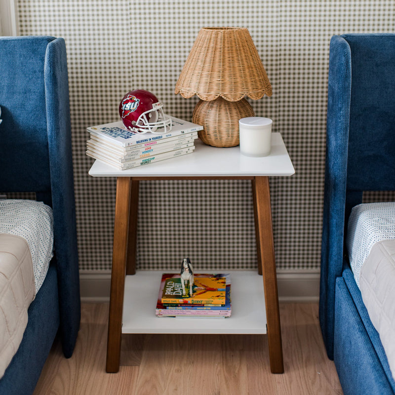 Mid-century modern bedside table with books, a rattan lamp, and a small football helmet, flanked by blue upholstered beds.