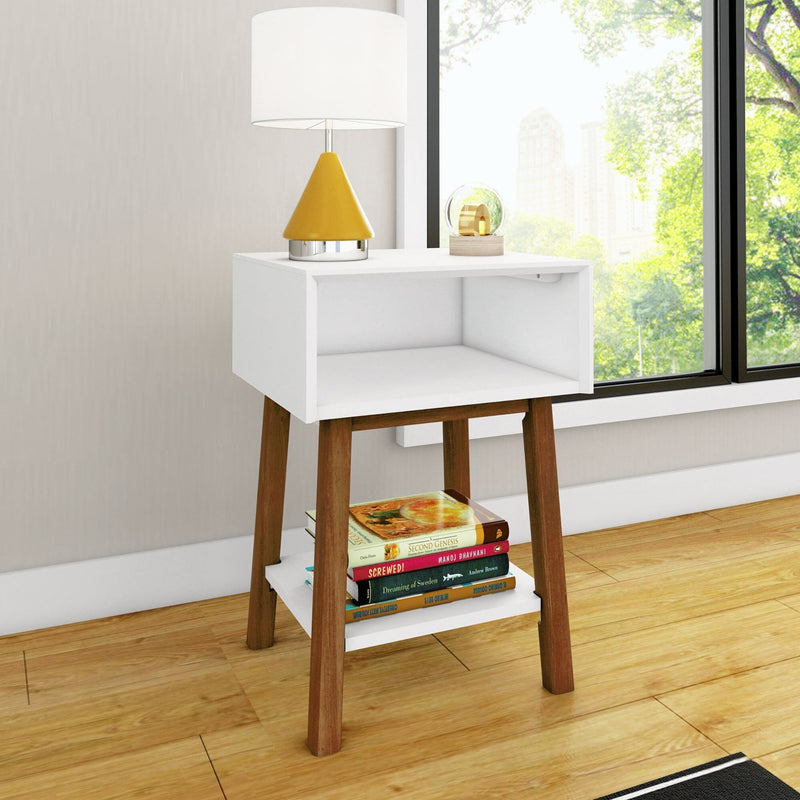 Modern white nightstand with wooden legs and open cubby, lower shelf stacked with books, yellow table lamp and glass orb on top, by a large window overlooking a city park on hardwood floor