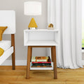 Minimalist mid-century modern white bedside table with wooden legs, yellow cone table lamp, glass orb decor and stacked books on a hardwood bedroom floor with white curtains