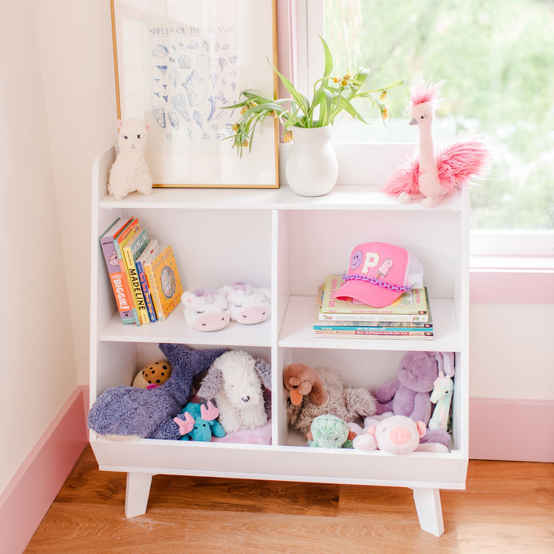 Child's bookshelf with stuffed animals, books, unicorn slippers, and a pink hat in a bright room.