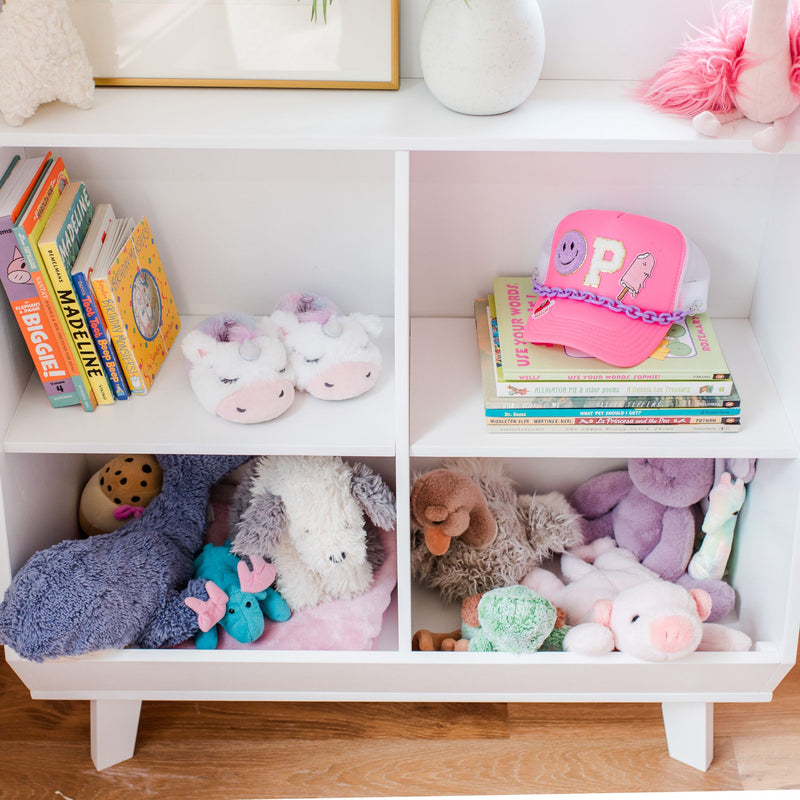 Kids' bookshelf with colorful books, pink cap, unicorn slippers, and plush toys on a white shelf.