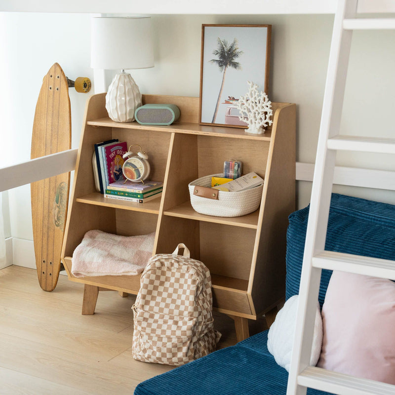 Kids bedroom wooden cubby storage shelf in a coastal-themed room holding books, alarm clock, lamp, woven basket, coral decor, checkerboard backpack and longboard