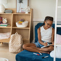 Young girl sitting cross-legged on a blue floor cushion in a cozy bedroom, drawing in a notebook with markers; checkered backpack and wooden shelf with books and lamp—back-to-school study scene.