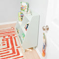 Mint-green angled children's bookshelf and toy organizer in a bright nursery or playroom, holding colorful picture books, plush toys and a toy train beside a white door and red geometric rug