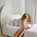 Girl playing with a plush doll on a neutral boho nursery canopy bed with quilted bedding, tassel pillows and a wooden balance board