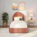 Modern bedroom with coral upholstered round headboard, yellow bedding and blue flower pillow, abstract framed wall art, wicker potted plant and wooden nightstand with lamp