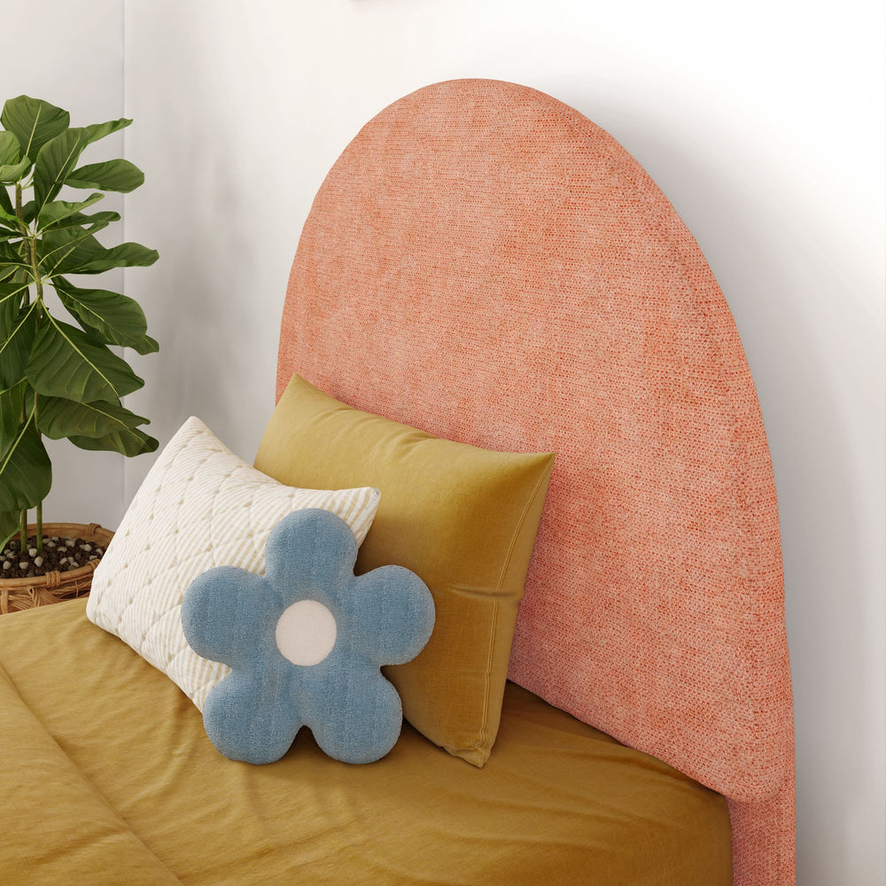 Stylish bed with pink fabric headboard, mustard bedding, and decorative pillows including a blue flower-shaped cushion, beside a leafy green plant.