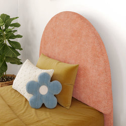 Stylish bed with pink fabric headboard, mustard bedding, and decorative pillows including a blue flower-shaped cushion, beside a leafy green plant.