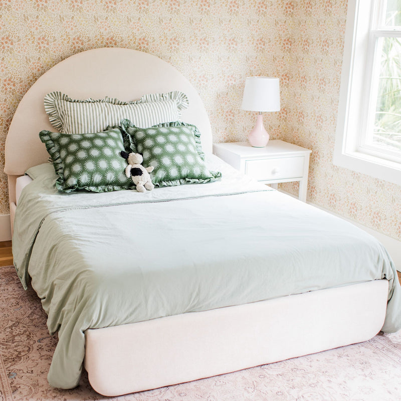Pastel floral wallpaper bedroom with arched upholstered headboard, mint bedding, green patterned pillows, white nightstand and pink lamp