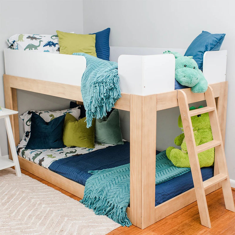 Light wood twin-over-twin kids bunk bed with white safety rails and ladder, teal and navy bedding, dinosaur-print sheets and green plush toys in a children's bedroom