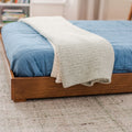 Cozy bedroom close-up of a wooden platform bed with a blue quilted comforter and cream sherpa throw blanket folded at the foot on a neutral textured rug, modern minimalist bedroom decor