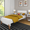 Modern minimalist bedroom with mustard yellow quilted bedding on a white platform bed, white bedside tables with yellow table lamps, abstract framed wall art, black-and-white striped rug and large window with leafy green view