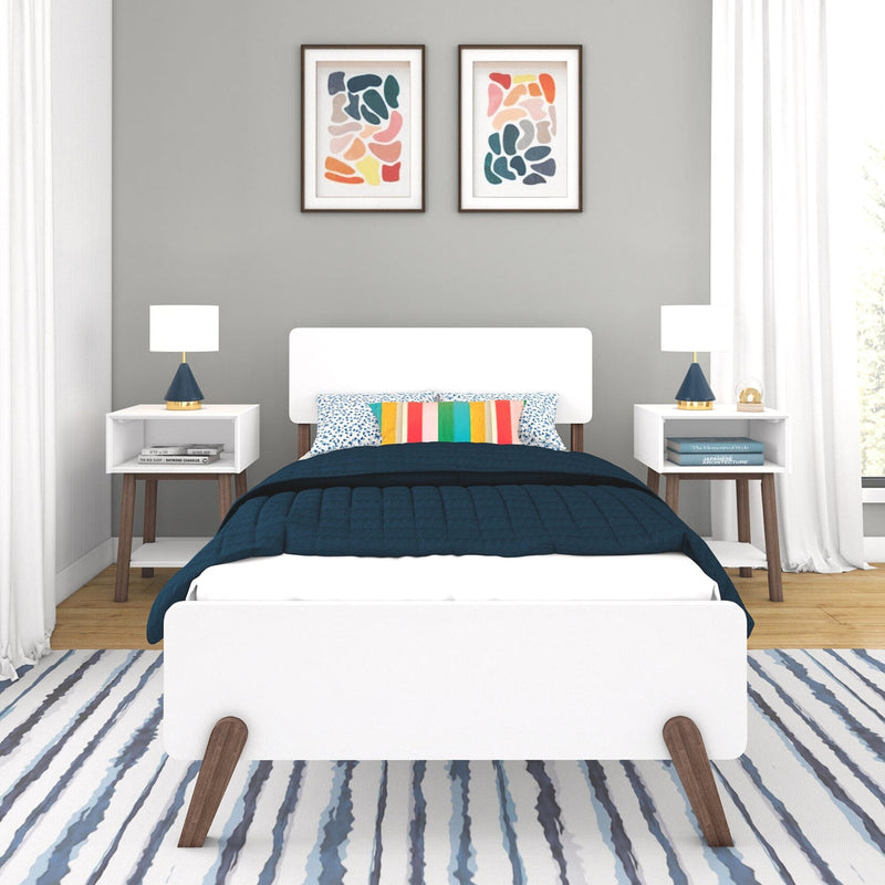 Modern bedroom with white bed, colorful striped pillows, navy quilt, abstract wall art, and white bedside tables with lamps.