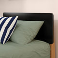 Close-up of modern black headboard with sage green bedding and navy-and-white striped pillow on wooden bed frame, minimalist bedroom decor