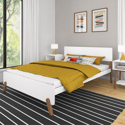 Modern white platform bed with mustard yellow quilt and patterned pillows on black-and-white striped rug, flanked by mid-century nightstands and lamps beside a large window and framed abstract art