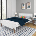 Modern minimalist bedroom interior with white platform bed on angled wooden legs, navy quilted bedding, colorful striped accent pillow, white nightstands with lamps, abstract wall art, large window and blue-striped area rug