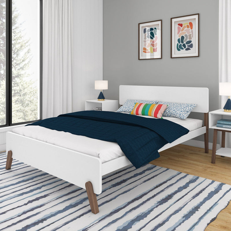 Modern minimalist bedroom interior with white platform bed on angled wooden legs, navy quilted bedding, colorful striped accent pillow, white nightstands with lamps, abstract wall art, large window and blue-striped area rug