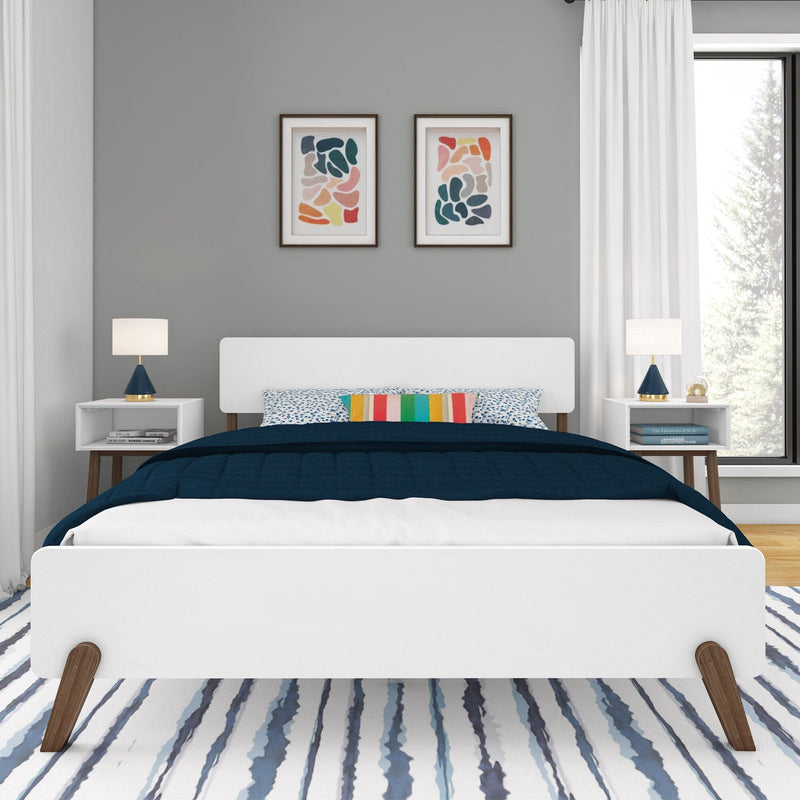 Modern mid-century bedroom with white platform bed, navy quilt, colorful striped accent pillow, matching nightstands and lamps, abstract wall art, gray accent wall, and blue-striped rug by a large window