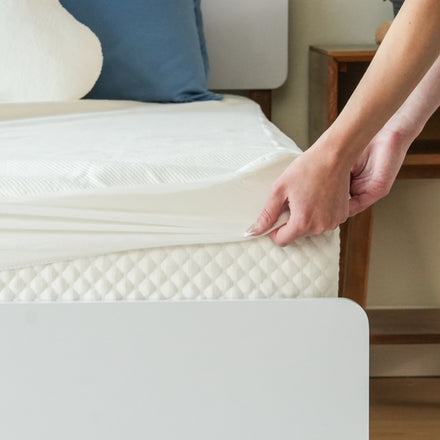 White fitted mattress protector being pulled onto a mattress on a modern bed with a blue pillow