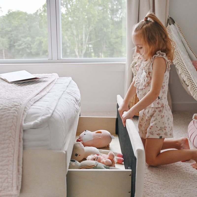 Little girl kneeling by an under-bed storage drawer, organizing plush toys in a bright neutral children's bedroom