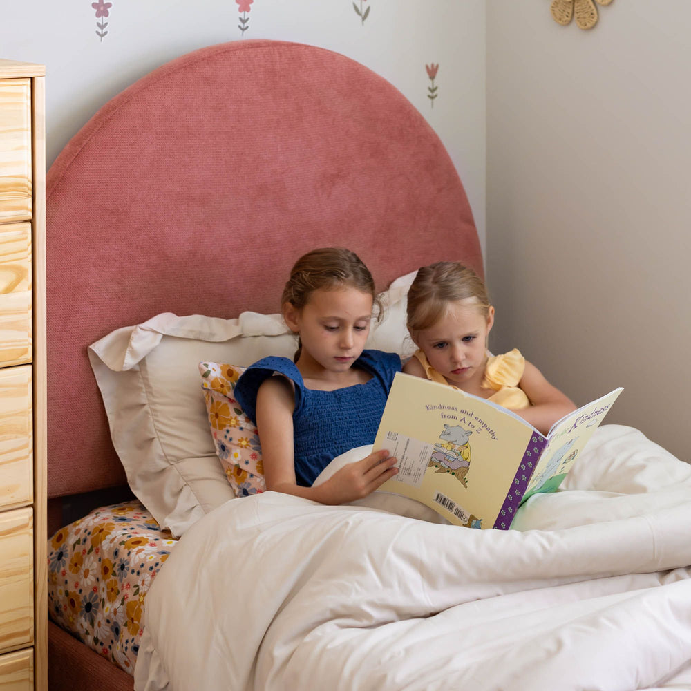 Two children reading a book together in bed with a pink headboard and floral pillow.