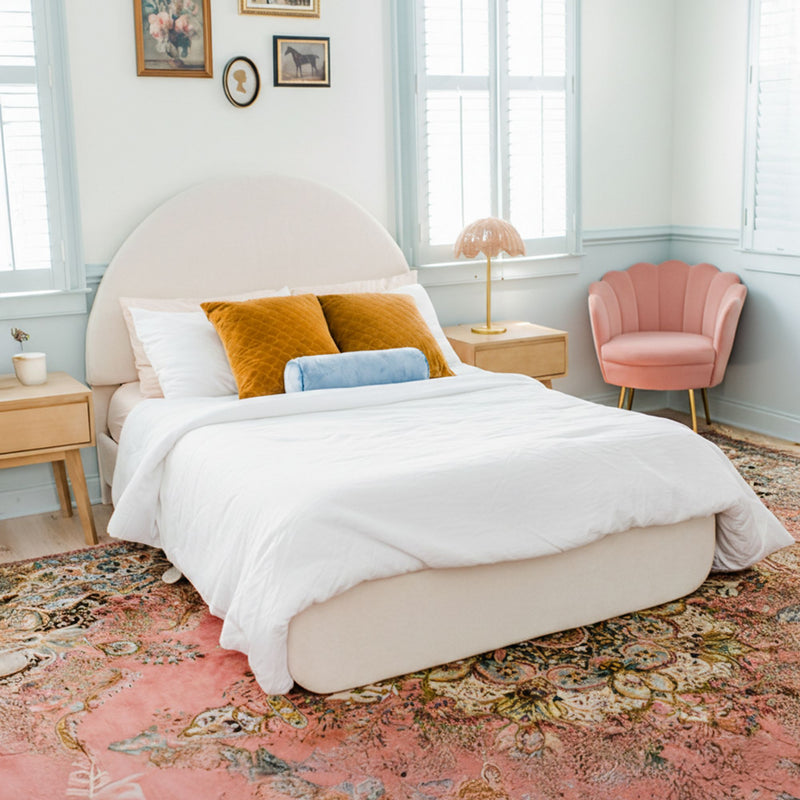 Sunlit modern boho bedroom with arched upholstered bed, white bedding, mustard velvet pillows and blue bolster, pink floral area rug, scalloped pink accent chair, natural wood nightstands and gold lamp