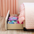 Under-bed storage drawer filled with pink dress-up costumes, tiaras and toys beside a pink quilted bed in a girl's bedroom