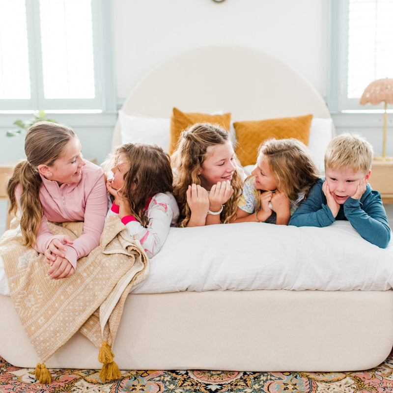 Children laughing and talking while lying on a bed in a brightly lit room.
