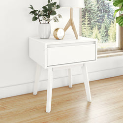 White minimalist Scandinavian bedside table with single drawer, tapered legs, potted greenery, wooden tripod lamp and gold alarm clock on hardwood floor beside window overlooking pine forest