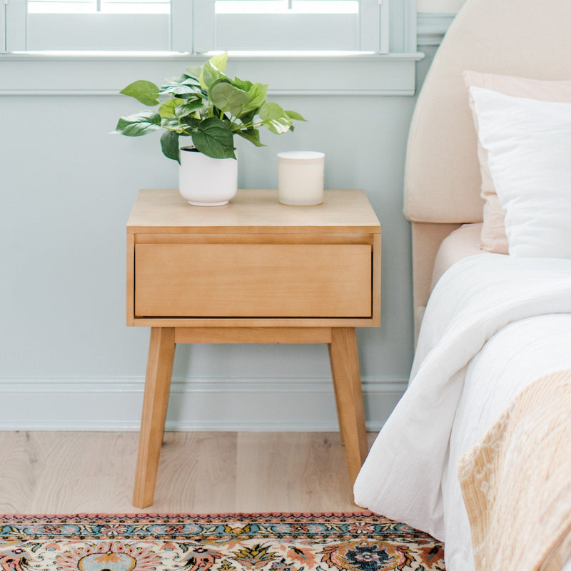 Light wood mid-century bedside table with drawer beside upholstered bed, potted green plant and candle on top, pale blue wall and patterned area rug, modern minimalist bedroom decor