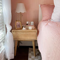 Cozy pink bedroom featuring a wooden mid-century bedside table with a scalloped white lamp, gold frame, stacked books, small vase of flowers and blush quilted bedding