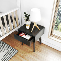 Modern black nightstand with open drawer, books, table lamp, small clock, and decorative plant, next to a wooden crib in a bright room.