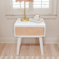 Modern mid-century white and natural wood nightstand with angled legs, brass lamp with pink shade, children's book and small white portable speaker on hardwood floor by shuttered window, nursery bedside decor