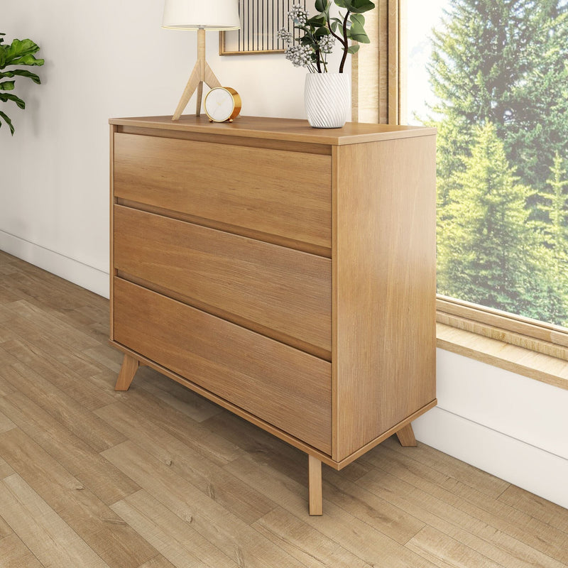 Mid-century modern oak three-drawer dresser on hardwood floor by sunlit window, styled with lamp, clock and vase — minimalist bedroom storage
