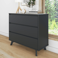 Modern matte-black three-drawer dresser with angled mid-century legs, styled with lamp, clock, books and potted plant beside a window overlooking green trees