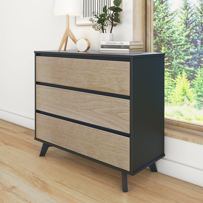 Mid-century modern three-drawer dresser with matte black frame and natural wood drawer fronts on angled legs, bedroom storage console styled with lamp, clock and potted plant