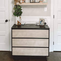 Modern three-drawer black and light-wood dresser in neutral nursery with potted plant, stuffed bear on shelf, baby monitor and framed photo — nursery storage and decor