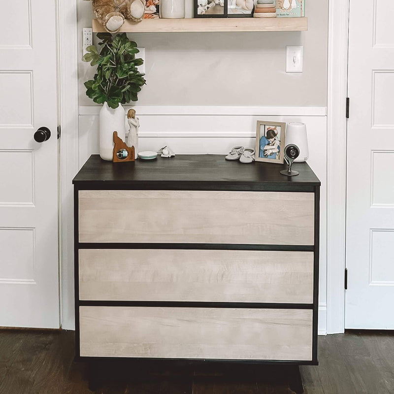 Modern three-drawer black and light-wood dresser in neutral nursery with potted plant, stuffed bear on shelf, baby monitor and framed photo — nursery storage and decor