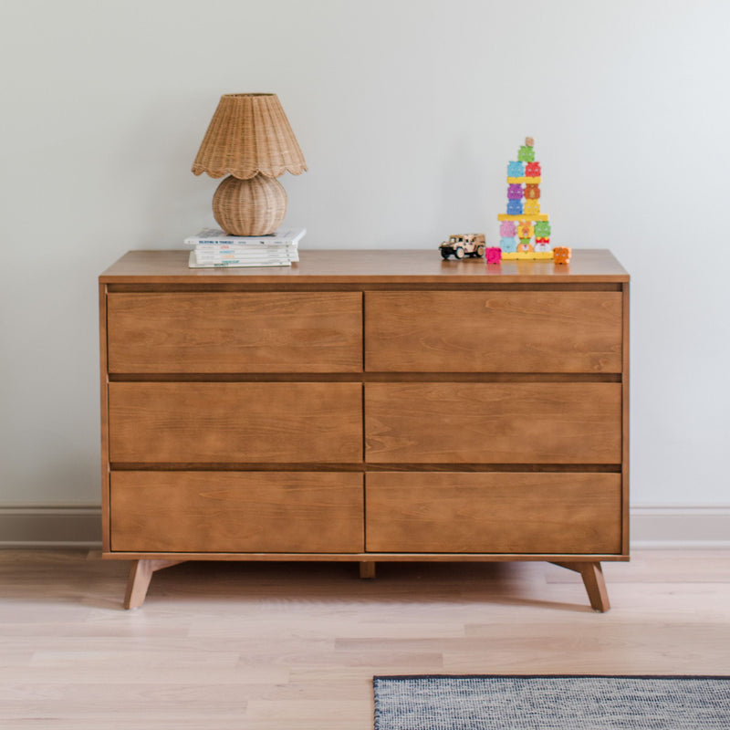 Mid-century modern six-drawer wood dresser topped with a woven rattan lamp, colorful children's stacking toy and toy car in a minimalist nursery bedroom
