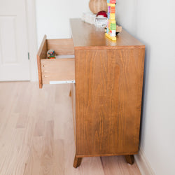 Mid-century wooden dresser with an open drawer and children's toys on top in a bright nursery with light hardwood floors