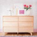 Light natural wood six-drawer mid-century modern dresser on hardwood floor styled with a white ceramic lamp, framed pastel artwork and pink peonies in a white vase against wave-patterned pink wallpaper