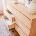 Wide light wood dresser with two open drawers, ceramic vases on top, pink patterned rug and hardwood floor in a minimalist bedroom.