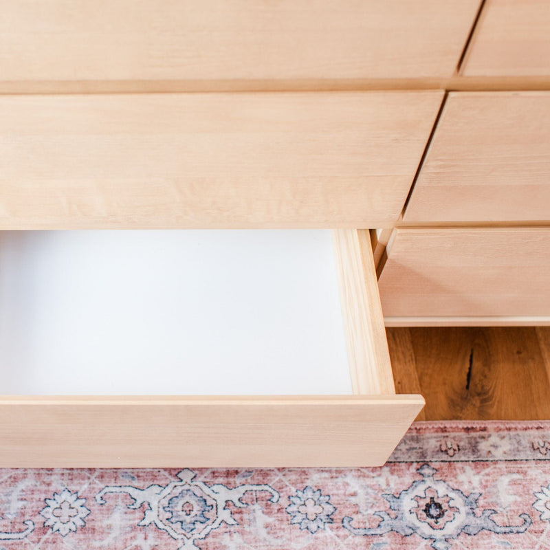 Open empty light wood dresser drawer in minimalist bedroom with white-lined interior, patterned area rug and hardwood floor