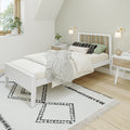 Minimalist Scandinavian bedroom with white wooden bed, layered neutral bedding and textured throw, black-and-white patterned area rug, wooden nightstand with alarm clock and plant, wall sconces and natural window light