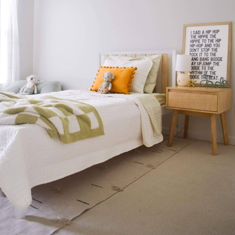 Cozy children's bedroom with white bedding, green-patterned blanket, decorative pillows, and plush toys beside a wooden nightstand.