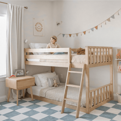 Smiling child on top of light wood kids bunk bed in neutral Scandinavian-style bedroom with ladder, bunting, stuffed animals and bedside table