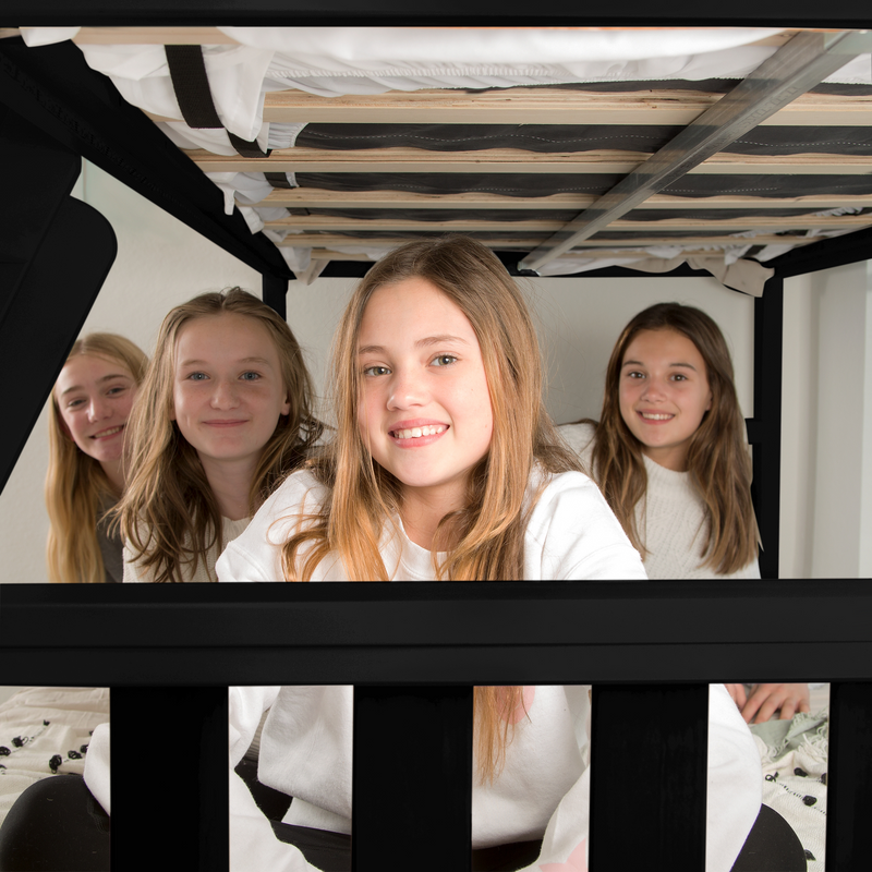 Four smiling girls having a sleepover under a black metal bunk bed in a bright, cozy bedroom